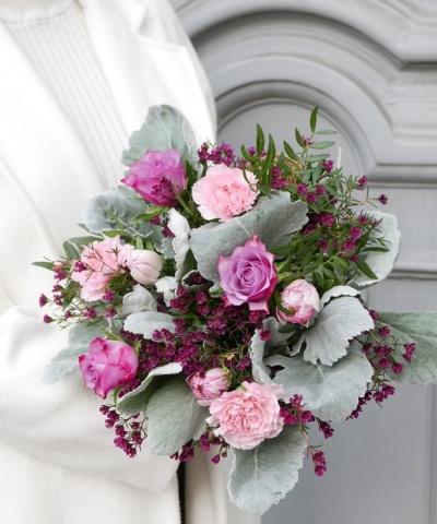 Buquê de flores rosa e lilás com folhagem prateada segurado por pessoa com casaco branco