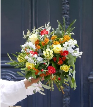 Buquê colorido de flores e folhas verdes segurado por mão com manga branca em frente a porta azul.