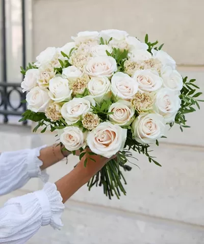 Buquê de rosas brancas com flores creme e folhagem verde, mãos em manga branca segurando