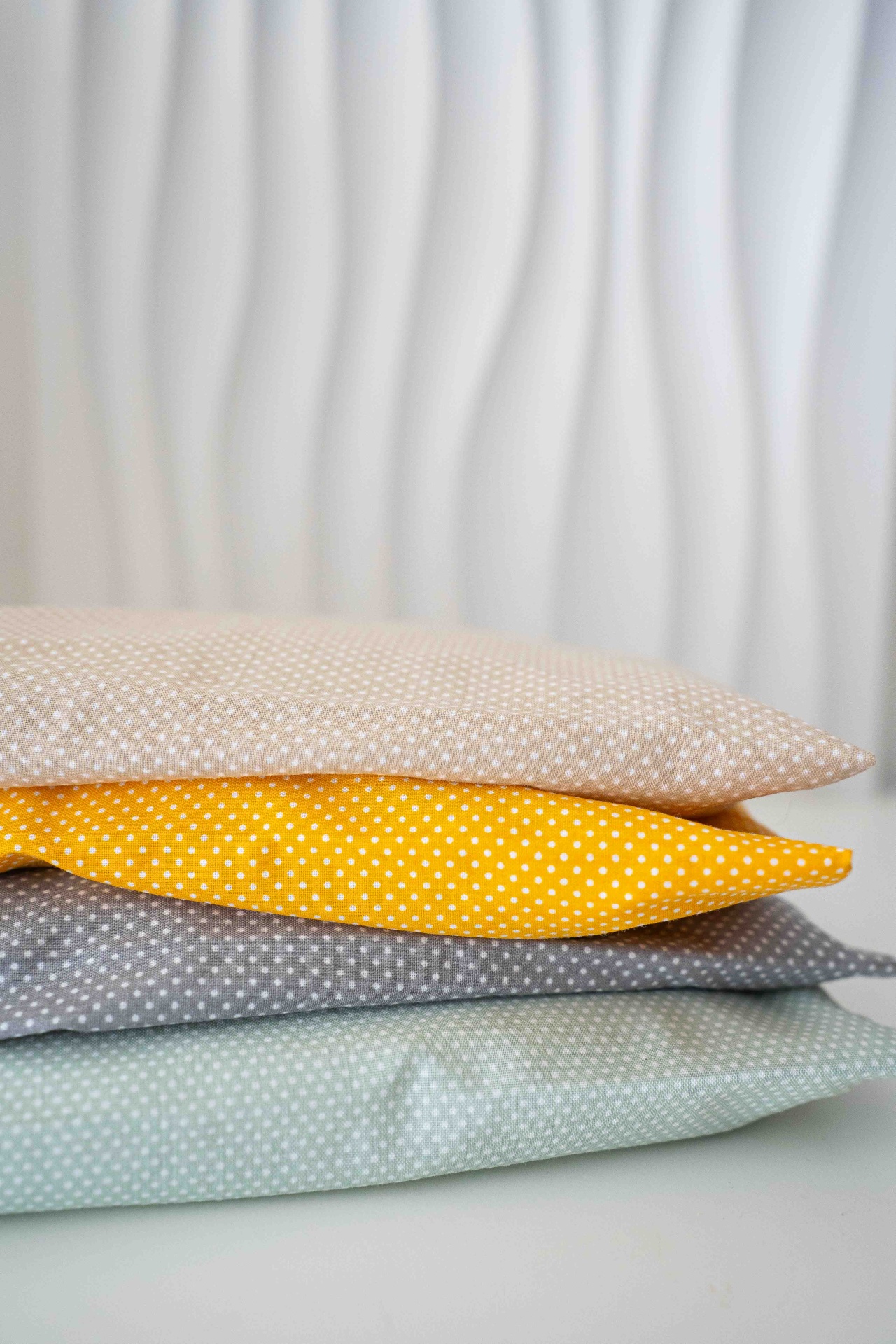 Almofada Terapêutica Três almofadas empilhadas com padrão de pequenos pontos brancos em fundos bege, amarelo e cinza.