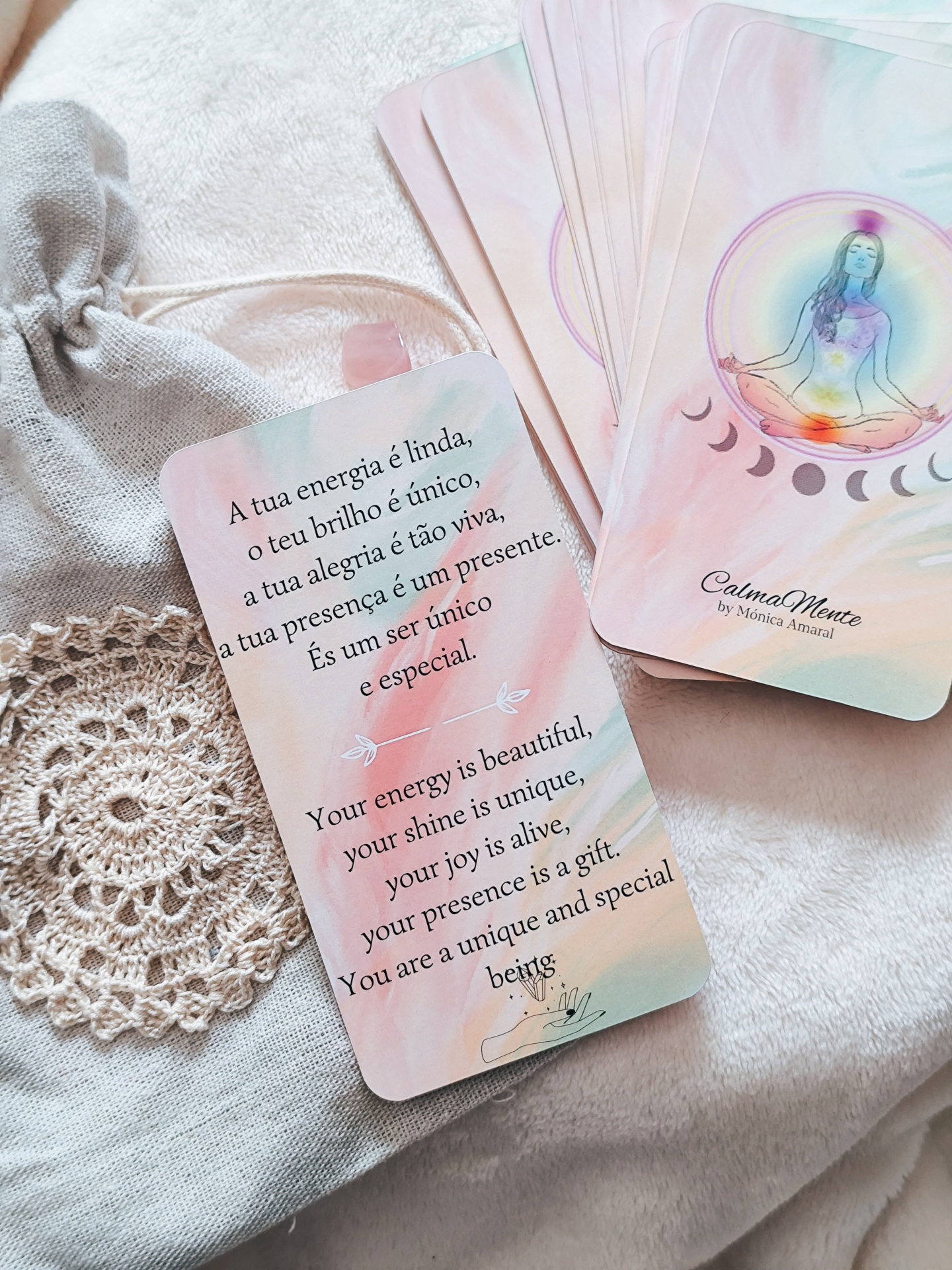 Oráculo CalmaMente Cartas de mensagens bilingues em tons pastel sobre energia e meditação, com saco tecido cinzento e renda bege no fundo