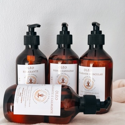 Quatro frascos âmbar com óleo relaxante com diferentes rótulos em português dispostos sobre superfície clara