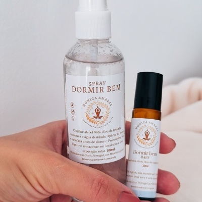 Frascos de spray e roll-on 'Dormir Bem' da marca Mônica Amaral numa mão, com fundo branco desfocado.