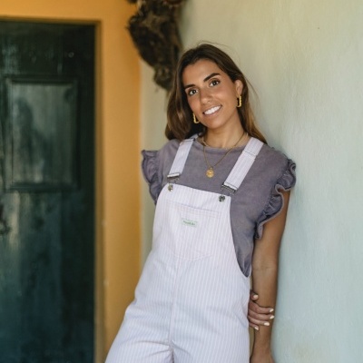 Macacão rosa claro riscado com t-shirt lilás com folhos, usado por mulher sorridente em ambiente interior