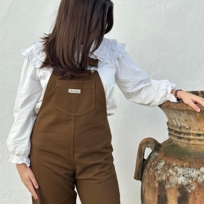 Macacão castanho com bolso frontal e etiqueta MonKiki e camisa branca com gola de babados ao lado de vaso cerâmico grande.