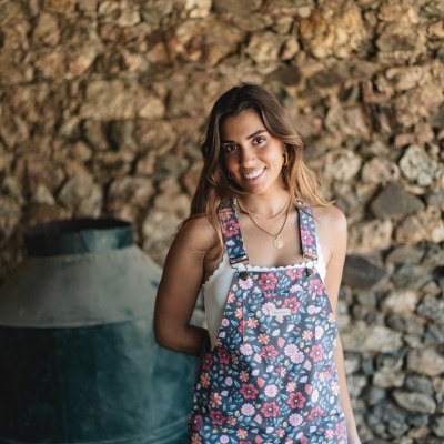 Mulher com jardineira floral colorida e blusa branca em frente a parede de pedra