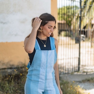 Mulher com macacão azul claro e camisola preta ao ar livre junto a portão de metal e flores amarelas no chão