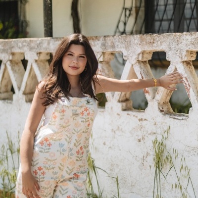 Macacão branco floral com etiqueta Moniká usado por modelo junto a muro e vegetação