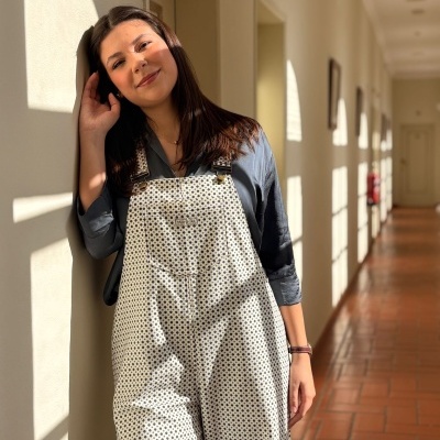 Mulher com macacão branco de bolinhas pretas e camisa cinza-azulada, encostada na parede de corredor iluminado.