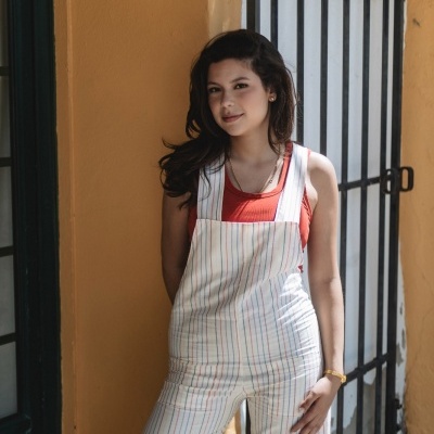 Mulher com macacão branco riscado e top vermelho junto a parede amarela.