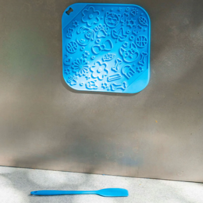 Tapete de silicone azul com relevos variados pendurado numa parede metálica com espátula azul à frente