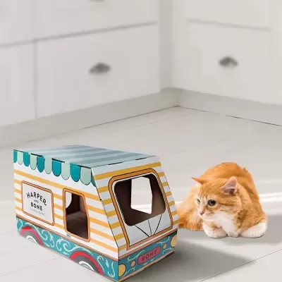 Abrigo de gato em forma de carrinha colorida de cartão numa cozinha branca com gato laranja ao lado