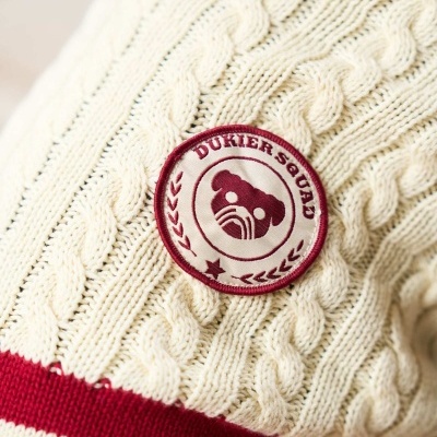 Suéter creme de malha com tranças e riscas vermelhas e emblema bordado com texto DUKIERSQUAD.
