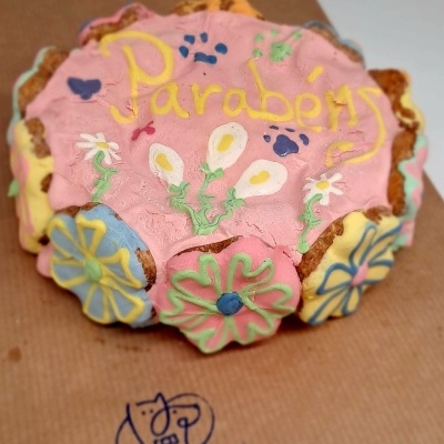 Bolo decorado com glacê rosa e flores coloridas com texto Parabéns sobre papel castanho com logotipo