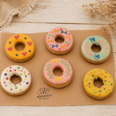 Biscoitos de cachorro coloridos em forma de donuts decorados, sobre tapete castanho com logo MORDOMIAS Pet Gourmet