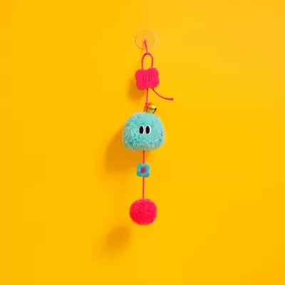 Chaveiro colorido pendurado com pompom azul, flores rosa e pompom vermelho num fundo amarelo