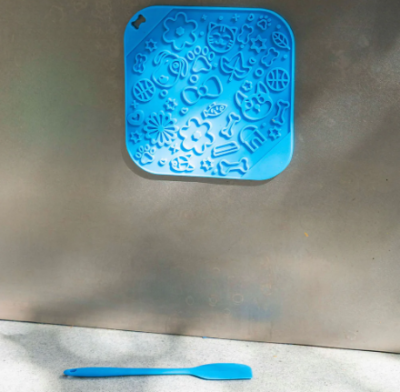 Tapete de silicone azul com relevos variados pendurado numa parede metálica com espátula azul à frente