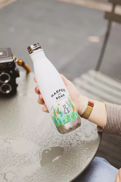 Garrafa térmica branca com decoração colorida e texto HARPER AND BONE na mão sobre mesa metálica molhada