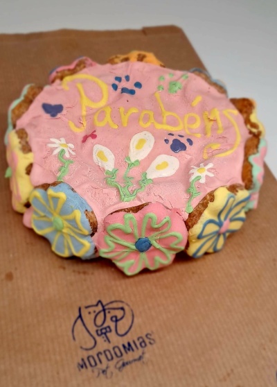 Bolo decorado com glacê rosa e flores coloridas com texto Parabéns sobre papel castanho com logotipo
