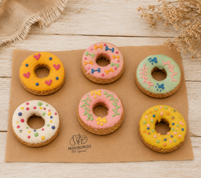 Biscoitos de cachorro coloridos em forma de donuts decorados, sobre tapete castanho com logo MORDOMIAS Pet Gourmet