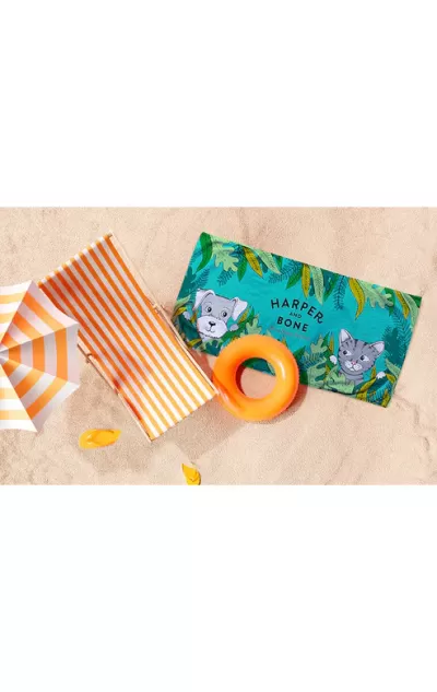Toalha de praia verde com cão e gato e texto HARPER AND BONE, colchão insuflável laranja, chapéu riscas brancas e laranja sobre areia