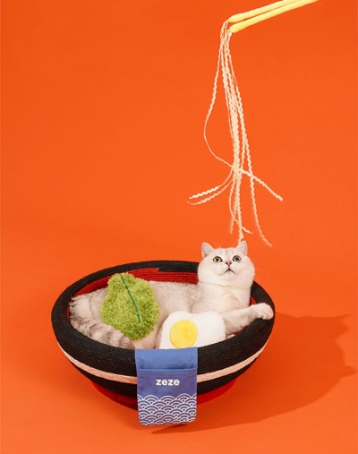 Gato branco numa cama redonda tricolor com brinquedos, fundo laranja.