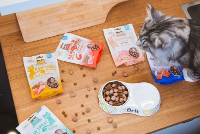 Petiscos para gatos Brit Raw Treat em vários sacos coloridos com gato a comer em bancada de madeira.