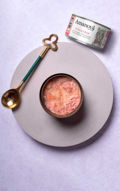 Lata de comida para animais Amanovã Tuna & Crab aberta junto a colher dourada com cabo verde sobre fundo claro