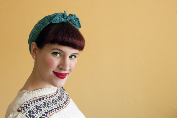 Mulher com bandana azul e camisola branca com padrão sobre fundo amarelo.