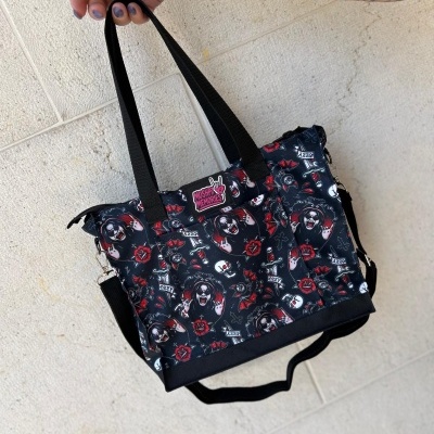 Black tote bag with dark-themed cartoon prints and text MOSHPIT MEMORIES held by tattooed hand
