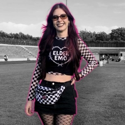 Young woman in ELDER EMO crop top, checkered long sleeves, black mini skirt, fishnet tights, checkered fanny pack, sunglasses, standing on grass in stadium.