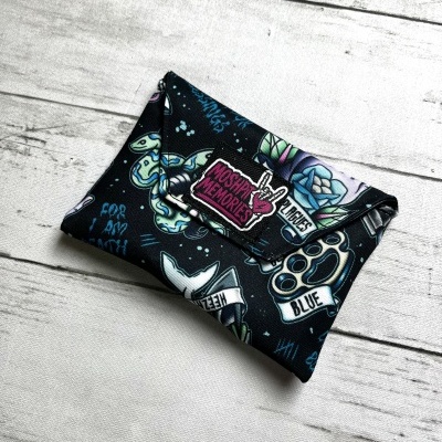 Folded black fabric pouch with tattoo designs and text MOSHPIT MEMORIES on white wooden surface