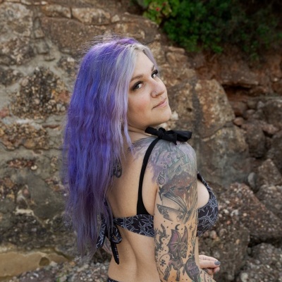 Woman in black patterned bikini with purple hair and tattoo sleeve near rocky wall