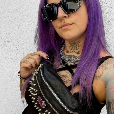 Black leather waist bag with silver spikes and patch, worn by person with purple hair and black sunglasses