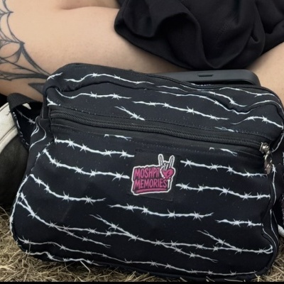 Black waist bag with white barbed wire pattern and MOSHPI MEMORIES patch on grass near tattooed leg and shoe