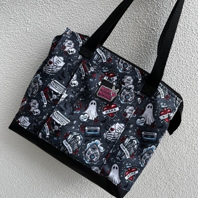 Black tote bag with tattoo-style print featuring skulls, ghosts, hearts, tombstones, and text labels.