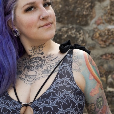 Black star-patterned bikini top, purple-haired woman with tattoos and sunglasses, stone wall background
