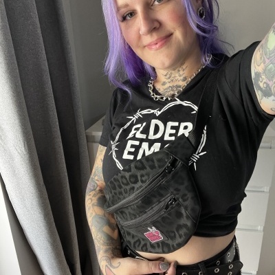 Person with purple hair wearing a black crossbody bag with leopard print and pink patch inside a room.