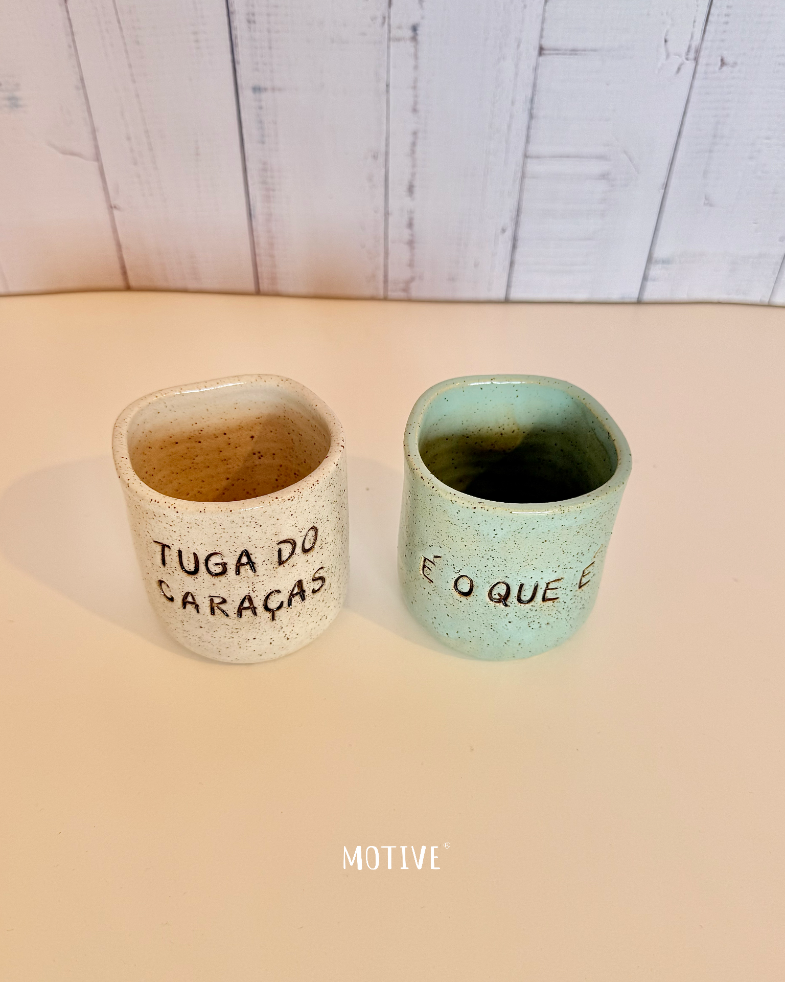 Dupla Copinhos TUGA DO CARAÇAS e É O QUE É Dois copos de cerâmica pontilhados, creme com texto e verde água com texto, sobre superfície clara e parede de madeira ao fundo.