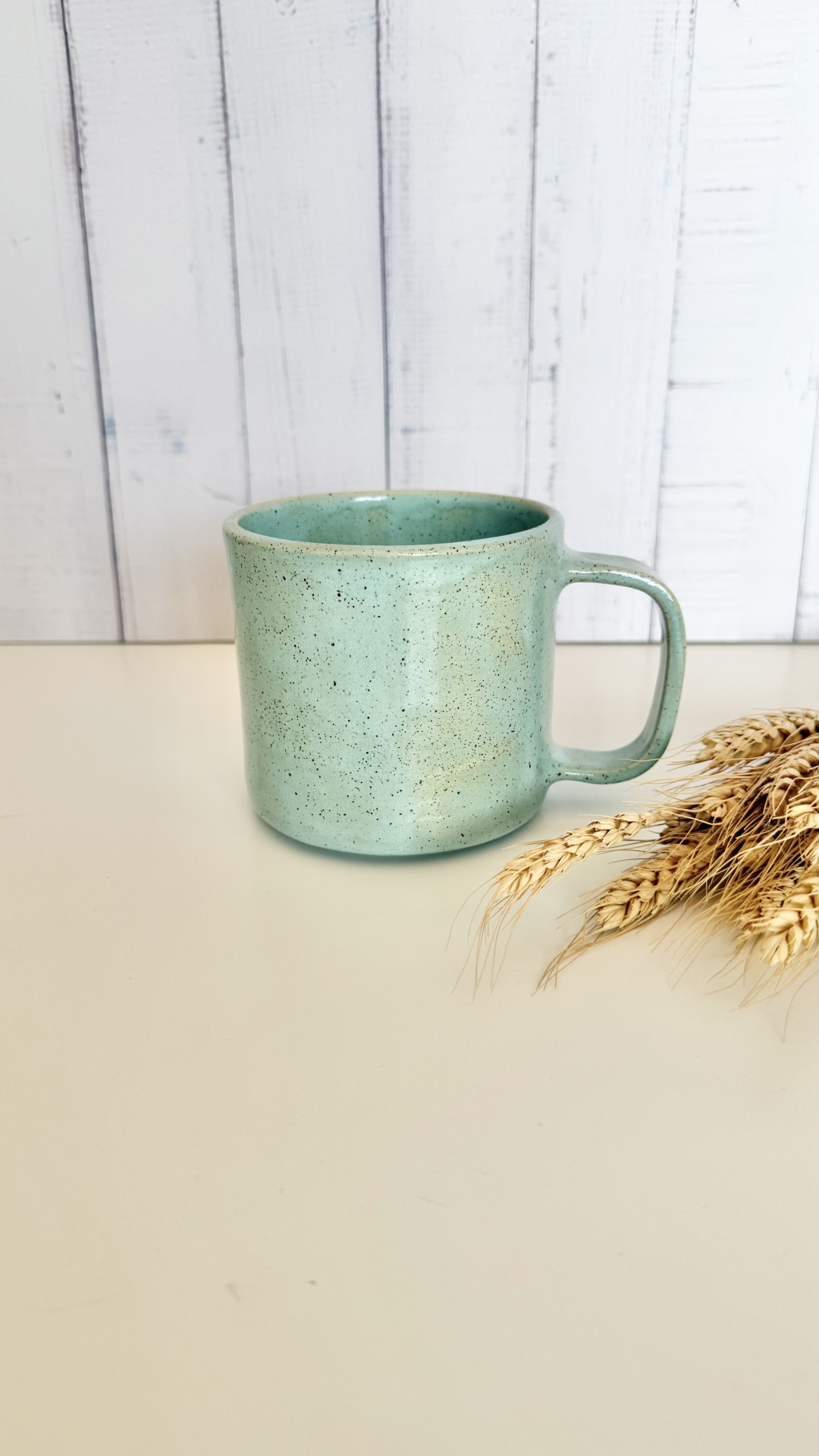 Caneca Calma Caralho Verde Caneca verde de cerâmica sobre bancada branca com espigas de trigo ao lado