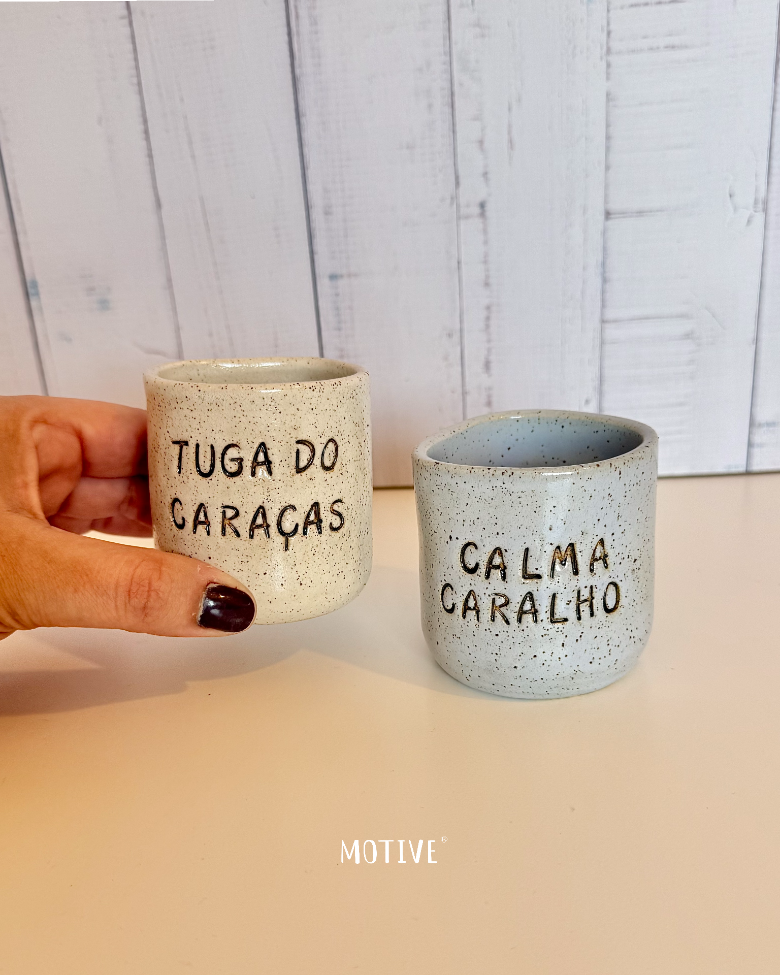 Dupla Copinho TUGA DO CARAÇAS e CALMA CARALHO Duas canecas de cerâmica com texto, uma creme e outra cinza, sobre superfície branca com fundo de madeira