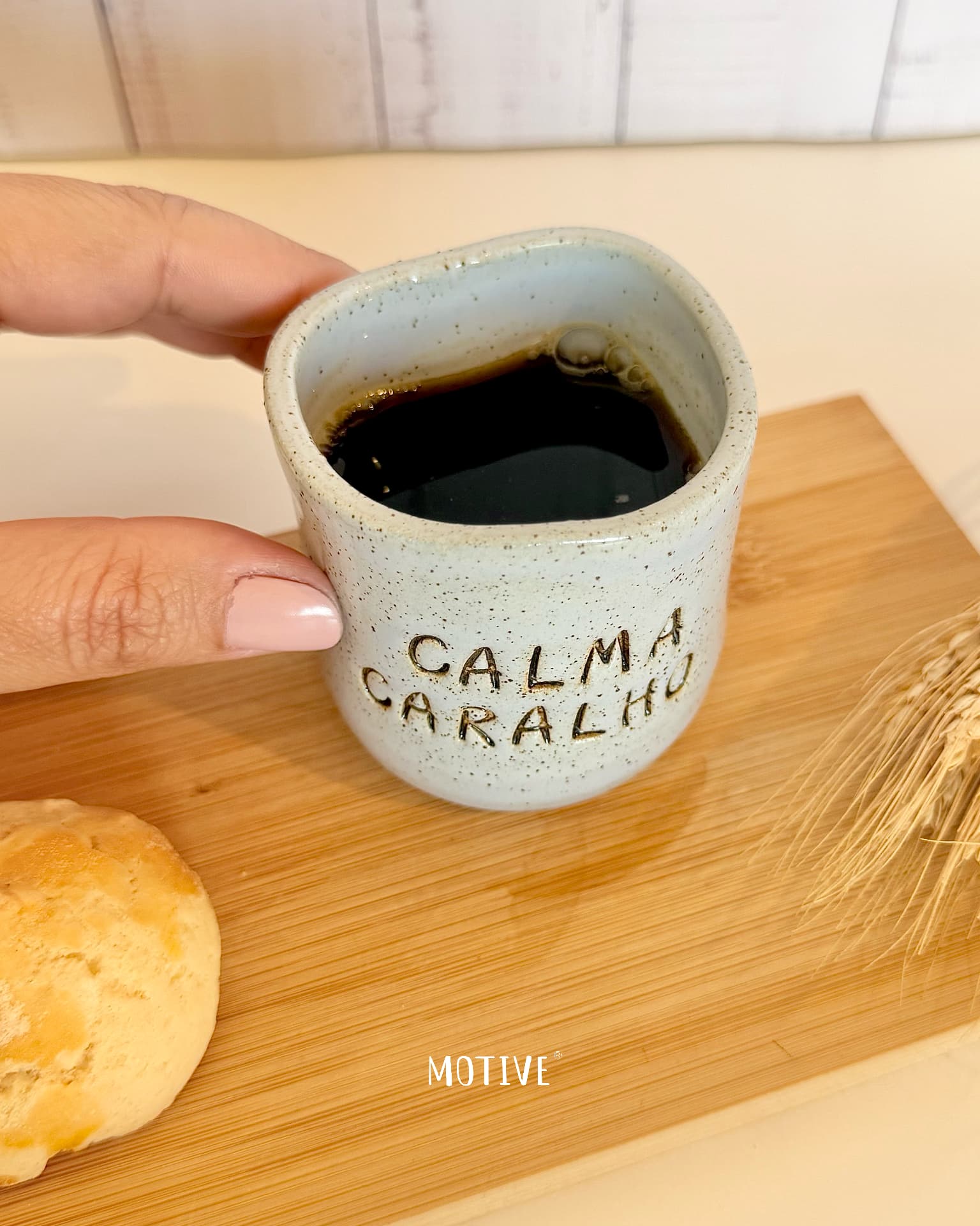 Copinho de Café CALMA CARALHO - Azul Celeste Caneca de cerâmica bege com texto CALMA CARALHO, cheia de líquido escuro, sobre tábua de madeira com bolacha e espigas de trigo