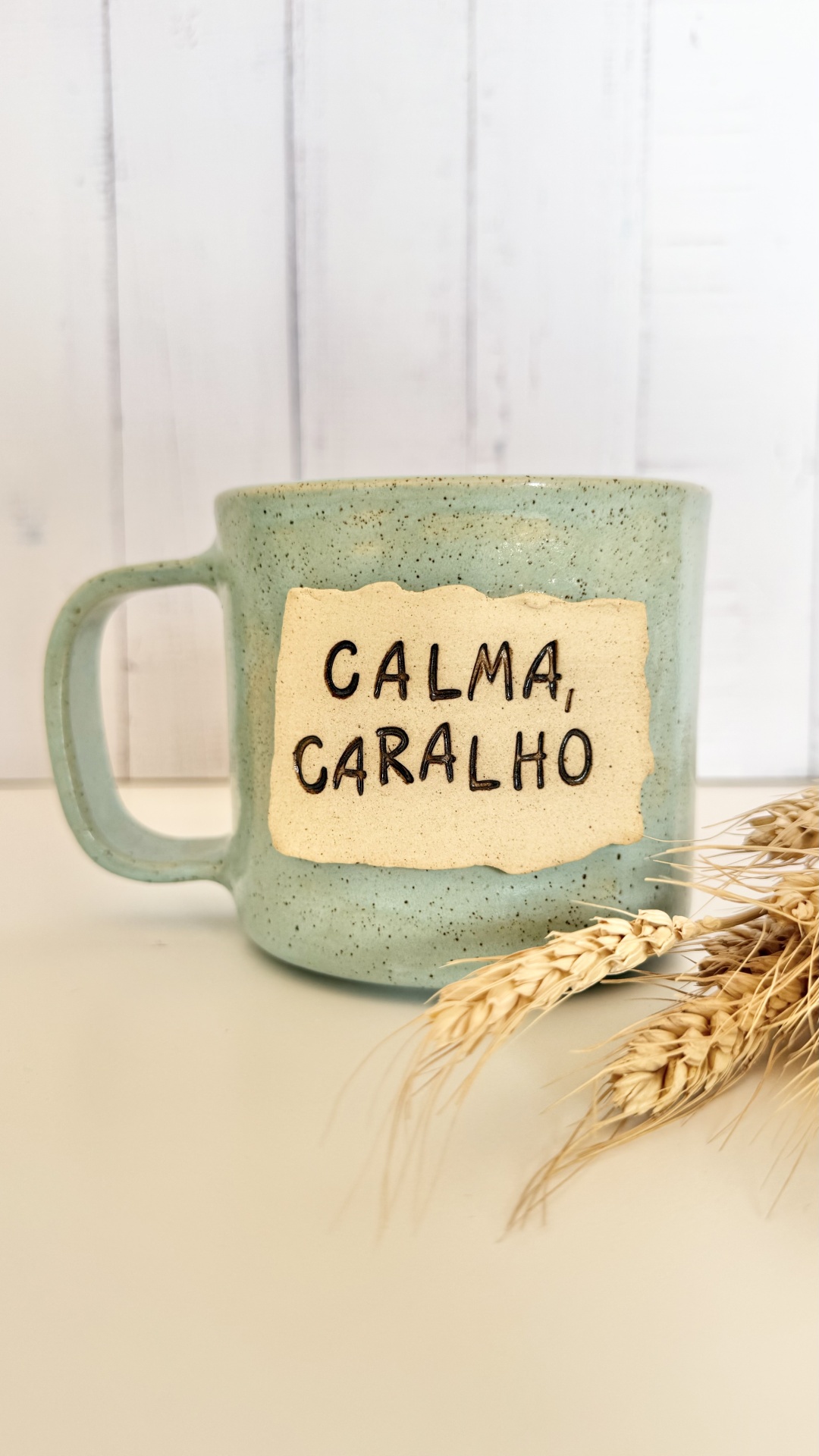 Caneca Calma Caralho Verde Caneca verde cerâmica com placa bege e texto CALMA, CARALHO ao lado de espigas de trigo