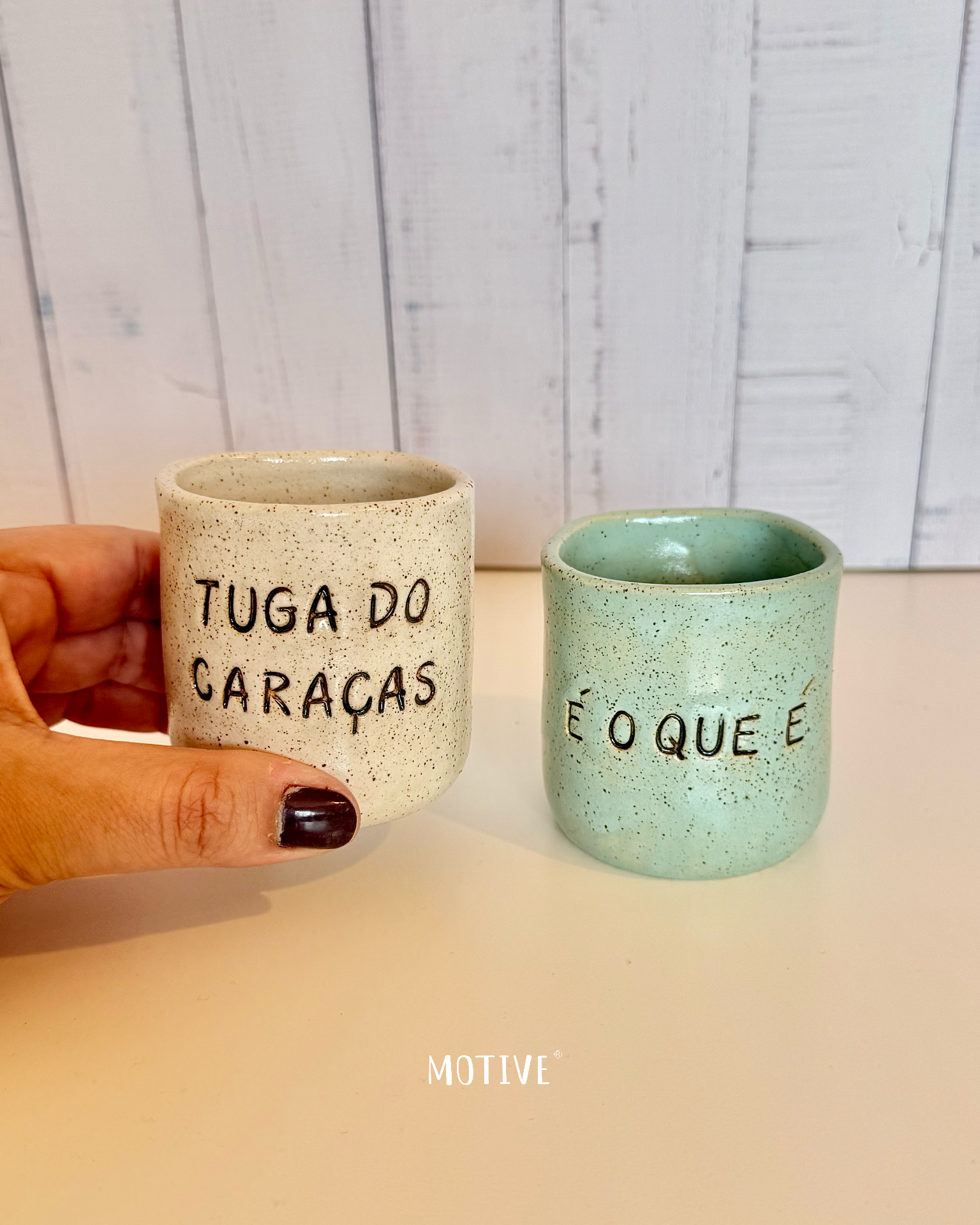 Dupla Copinhos TUGA DO CARAÇAS e É O QUE É Dois vasos em cerâmica com textos em português, um bege e outro verde água, sobre superfície branca e fundo de madeira.