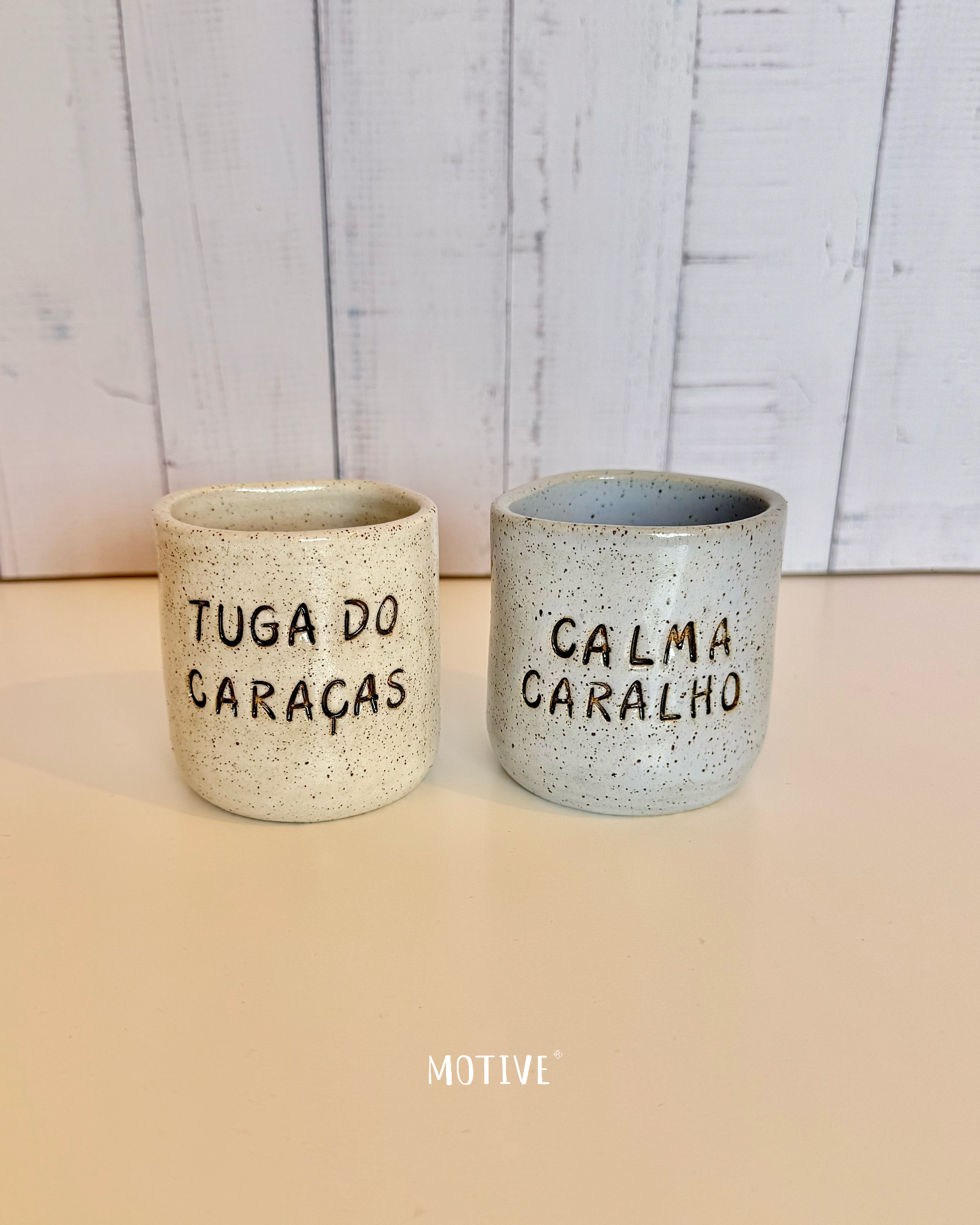 Dupla Copinho TUGA DO CARAÇAS e CALMA CARALHO Duas canecas de cerâmica salpicada com textos em preto sobre fundo branco de madeira.