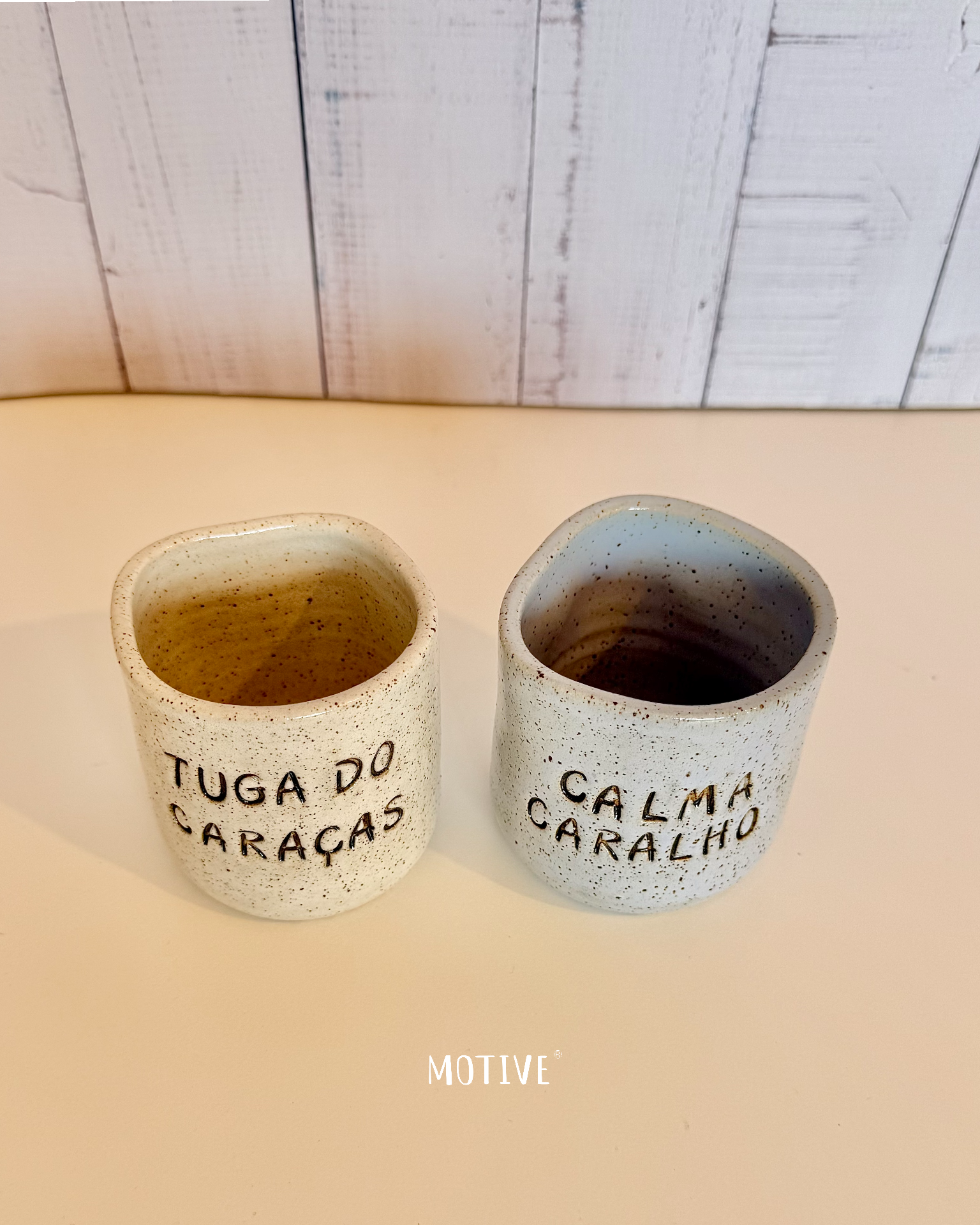 Dupla Copinho TUGA DO CARAÇAS e CALMA CARALHO Dois copos cerâmicos com textos em português sobre superfície branca e fundo madeira