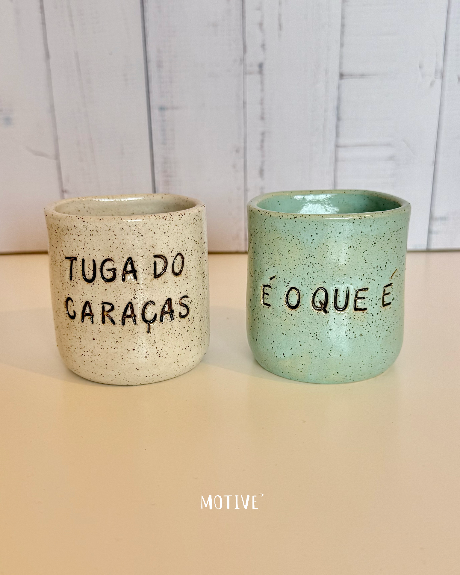 Dupla Copinhos TUGA DO CARAÇAS e É O QUE É Dois copos de cerâmica com inscrições em português sobre mesa clara e fundo de madeira branca.