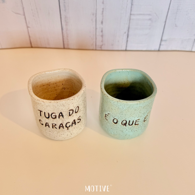 Dois copos de cerâmica pontilhados, creme com texto e verde água com texto, sobre superfície clara e parede de madeira ao fundo.