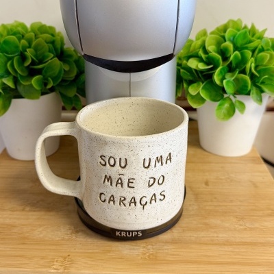 Caneca de cerâmica bege com texto em relevo e base preta KRUPS entre plantas verdes