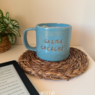 Caneca de cerâmica azul com texto CALMA, CARALHO sobre descanso de copo de fibras naturais